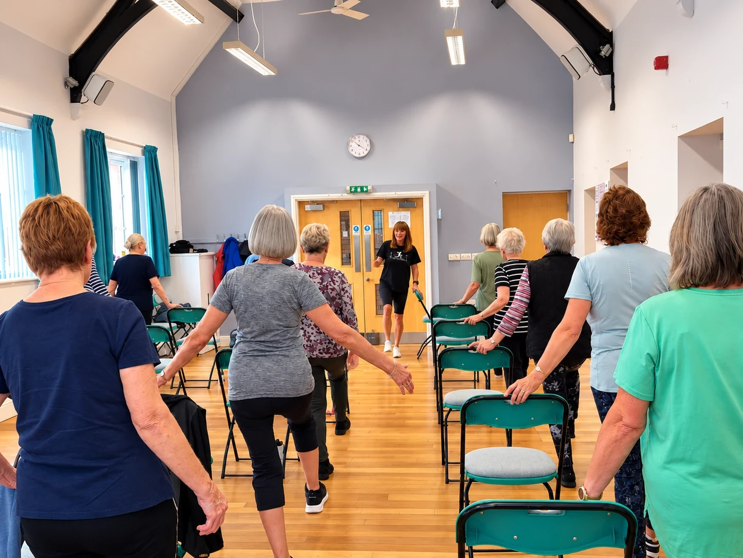 Vikki leading a chair-based class in a village hall, with members holding the backs of their chairs for a balance exercise